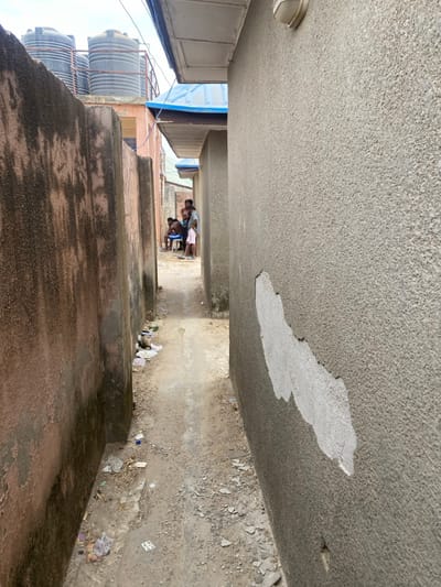 An image depicts a narrow unpaved alley in the city of Katcha, Nigeria, extending forward between two multi-story buildings under a hazy sky. The left wall features weathered reddish-brown rendering, while the right wall is light grey textured stucco with large sections of render peeled away, revealing a lighter subsurface. The unpaved ground consists of dirt and scattered litter, with a slightly raised central path of compacted earth or worn concrete. Approximately five individuals are gathered in the mid-ground of the alley: a shirtless male is seated on a white stool, surrounded by standing adults and a young child in a light-colored outfit. In the background, above the structures, multiple black plastic water storage tanks are visible on a rooftop. Overhanging eaves extend from the buildings above both alley walls. A light fixture is mounted on the upper section of the right wall.