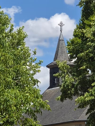 The image shows the upper portion of a church steeple with a cross on top, framed by green trees against a blue sky with white clouds. The steeple is covered in dark grey slates, and a small rectangular window with horizontal slats is visible on the side of the steeple. The cross is ornate and metallic. The surrounding trees are lush and leafy, with some of their branches obscuring parts of the church. The image appears to have been taken on a sunny day. The general architectural style and the presence of a church suggest this location could be in a European town, possibly within the city of Schengen, Luxembourg.