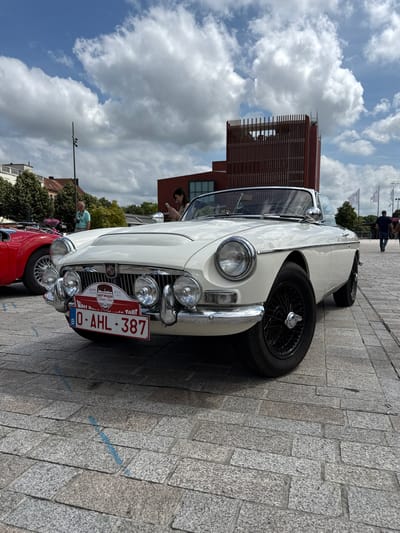 The image shows a white MG convertible parked on a cobblestone street in Bruges, Belgium. The car is the main subject, with its chrome accents and wire wheels clearly visible. A license plate reading "0-AHL-387" is attached to the front. Behind the car, a red car is partially visible. In the background, a modern building with a wooden facade stands under a cloudy sky. Several people are scattered around the area, some looking at the cars, others walking. The scene suggests a classic car event or tour, possibly a rally, given the "Classic Tour" text on the license plate. The time of day appears to be daytime, with bright, overcast conditions. The presence of cobblestones and the architectural style of the building in the background are strong indicators of the European location, specifically Bruges.