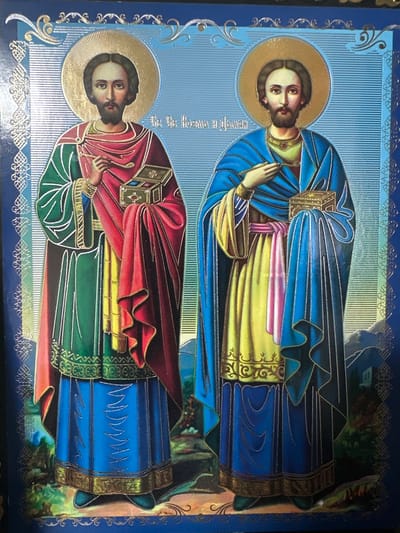 The image is an icon depicting two male figures with halos, dressed in robes. The figure on the left wears a green and red robe over a yellow undergarment and a blue skirt, holding a small ornate box in his right hand and a staff or brush in his left. The figure on the right wears a blue robe over a yellow undergarment with pink and purple accents, also holding a similar ornate box in his right hand and a staff or brush in his left. Both figures have beards and dark hair. They are depicted standing in a landscape with rolling hills and some distant structures. The background is a textured blue. Decorative gold borders frame the image. There is text in an unfamiliar script above the figures. The icon is likely an image of saints.

There is no information in the image to indicate it is located in the city of Marten, Bulgaria.