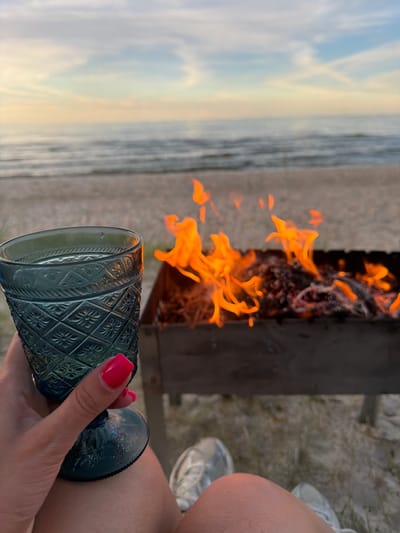 A person's hand with bright pink nail polish holds a dark blue, patterned glass. In the background, flames erupt from a grill or fire pit, with the fire casting a warm glow. To the left of the flames, a body of water, likely the sea, meets a sky filled with soft, diffused clouds, suggesting either dawn or dusk. The ground appears to be sandy, with some gravel. The overall scene evokes a sense of relaxation and enjoyment, possibly a beach gathering or a quiet moment by a fire near the coast. The light suggests it is late afternoon or early evening.