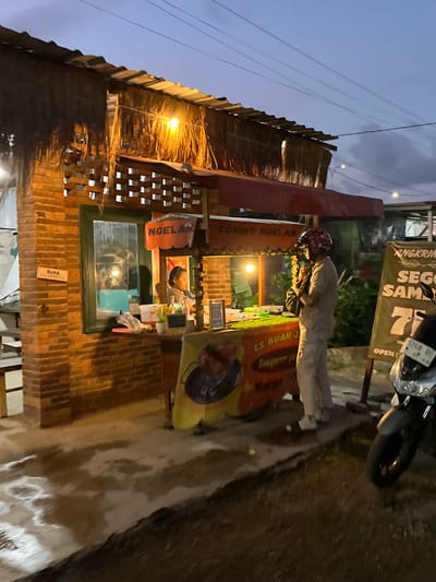 The image depicts an outdoor food stall in Kludan, Indonesia, during twilight. A brick structure with a thatched roof and red awning houses the stall. A person, wearing a light-colored t-shirt, pants, and a red helmet with white patterns, stands facing the stall, holding a bag and appearing to be in conversation with the vendor. The vendor, a young woman with dark hair, is visible behind a green-framed window, attending to the stall.
The stall itself is adorned with various signs, one in orange with "NGELAK" and "TOMBO NGELAK" written on it, and another in yellow and orange with text that includes "ES BUAN J" and "Harga". A motorcycle is parked to the right of the stall, with a sign in the background displaying "ANGKRINGAN SEGO SAMBEL 75". The ground in front of the stall is wet, with puddles and some white frothy residue. The overall atmosphere suggests a local eatery operating in the evening.
