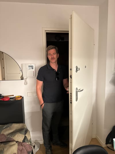 A man, with light brown hair and fair skin, is looking through an open doorway. He is wearing a black polo shirt and dark pants. His right arm is bent at the elbow, and he appears to be holding something small and metallic in his hand. He has a light tan and a casual expression.
The setting appears to be indoors, likely a hallway or entrance area, given the presence of a door and what looks like an intercom system on the wall. The door itself is white and has two distinct handles, one circular and one curved. The room is simply decorated, with plain white walls. A sliver of natural light is visible from an opening in the wall, suggesting it might be daytime.
There is a white intercom system mounted on the wall to the left of the man. It has a small screen and several buttons. Below the intercom, a portion of a large, arched mirror is visible. To the right of the intercom, there is a black cabinet with a camouflage jacket draped over it.
The overall impression is casual and everyday, with no specific event or activity being the primary focus. The man's pose and expression are relaxed, suggesting he is not in a hurry. The lighting and indoor setting point towards a typical day. The location context of Murcia, Spain, does not offer any specific visual cues in this image.