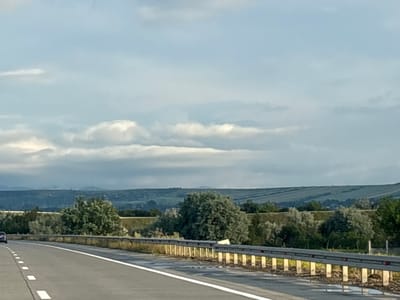 The image captures a view from a highway in Samtsevrisi, Georgia, looking across a landscape under a cloudy sky. The foreground features an asphalt road with white dashed lines on the left side, suggesting it is a multi-lane highway. A metal guardrail with concrete posts runs along the right edge of the road, with some vegetation and debris visible beneath it. Beyond the guardrail, the terrain is covered with trees and bushes of various sizes and densities, some appearing as large, rounded canopies. In the middle ground, rolling hills are visible, interspersed with what appear to be agricultural fields or terraced slopes, creating a textured pattern across the landscape. The background consists of hazy, dark hills or mountains shrouded in mist. The sky is predominantly pale blue with scattered white and gray clouds, some appearing soft and wispy, others more solid and layered. A dark-colored car is visible on the left side of the highway, moving away from the viewer. The overall impression is a rural highway scene during a day with overcast conditions.