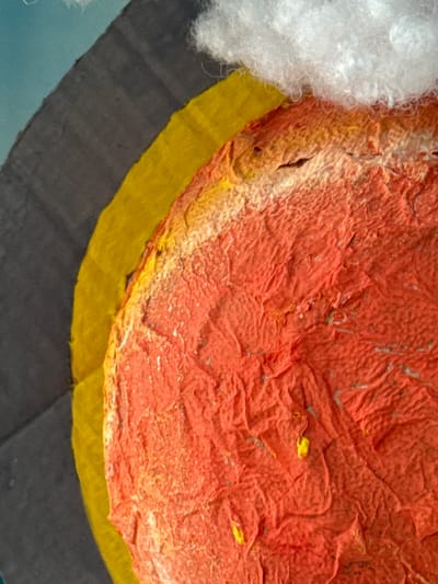 A close-up image shows layered materials in various colors. At the top is a textured, reddish-orange surface, potentially paint or paper. Below that is a band of yellow material. Beneath the yellow is a dark gray section, and on the left edge, there is a fluffy white substance. The scene does not depict the location of Ново-Никольское, Russia, or any specific city. There are no people or animals in this image, it is inanimate objects.