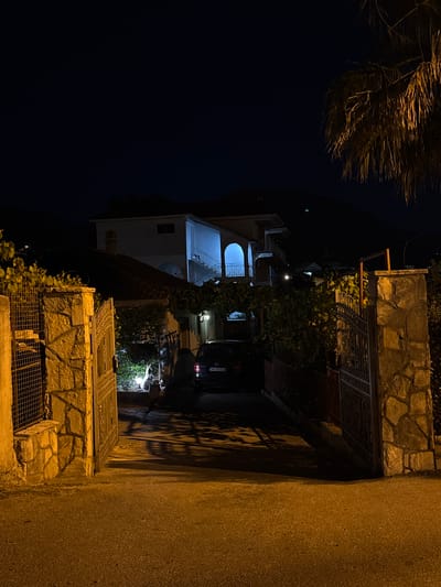 This is a night-time photograph taken from a street level view, looking towards an open gate. The gate is made of decorative metalwork and is flanked by stone pillars constructed from irregular, light-colored stones. Beyond the gate, a paved driveway leads to a multi-story building. The building has a white facade and features balconies with white railings and arched openings. Exterior lights illuminate the building, casting blue light on the upper floors and white light on the lower areas. A dark-colored car is parked in the driveway. Greenery, including vines and trees, is visible around the property, with a palm tree frond in the upper right corner of the image. Shadows from the gate and vegetation are cast across the driveway and road. The sky is dark blue, indicating night. The location is identified as Burtaiši, Montenegro.