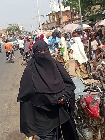 This image depicts a bustling street scene in Ogaminan, Nigeria, during the daytime under a clear sky.
In the foreground, a person is prominently featured, wearing a black niqab that covers their entire face except for their eyes, and a matching black abaya. They appear to be seated on a motorcycle, with a red motorcycle fender or fuel tank partially visible behind them. Their right hand, partially covered by the abaya, is holding onto something, and a wrist accessory is visible.
The background reveals a vibrant and crowded environment. To the left of the main subject, a busy market area is visible, teeming with people dressed in various attire, including traditional garments. Vendors are present, some behind stalls or displaying goods on the ground, with a light-colored umbrella offering shade. Further back, buildings with unpainted brick or concrete facades line the street. A white sign on a pole is discernible, though blurry, with some visible text fragments such as "AD", "OPO", and "N4,000".
To the right of the main subject and extending into the mid-ground, a busy street is packed with numerous individuals, many on motorcycles, suggesting heavy traffic and constant movement. The ground is paved, indicative of a developed road. The overall scene conveys a sense of everyday life and commerce in a lively West African urban setting.