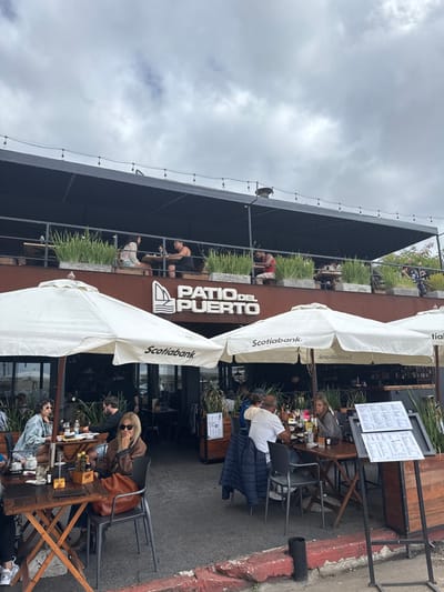 The image depicts an outdoor dining area of a multi-level restaurant, likely during daytime under an overcast sky. The scene is bustling with patrons seated at various tables.
The restaurant, prominently featuring the name "PATIO DEL PUERTO" on its facade, has both ground-level patio seating and an upper-level balcony area. On the ground level, several tables are arranged outdoors, shaded by large white umbrellas bearing the "Scotiabank" logo. People are seen socializing and dining; one woman with sunglasses and a brown jacket is seated at a table in the foreground, while further back, a man in a white shirt is engaged in conversation with others. Two large black menu boards stand near the seating area.
The upper level, separated by railings and planters filled with green foliage, also hosts several diners. Individuals are visible leaning against the railings and sitting at tables, enjoying the outdoor setting. The building's structure is dark, contrasting with the white umbrellas and the lighter cloudy sky.
The scene suggests a relaxed atmosphere, with people enjoying food and drinks. The ground-level patio borders a paved street or sidewalk, which has a distinct red curb. The sky is largely covered with clouds, indicating a cloudy or overcast day. The visible text includes "PATIO DEL PUERTO" on the building's sign and "Scotiabank" on the patio umbrellas. The location is contextualized as Punta del Este, Uruguay.
