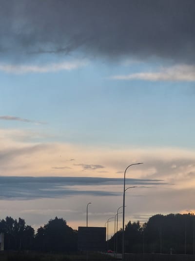 The image depicts a cloudy sky at dusk over a treelined landscape. Several streetlights are visible, illuminating a roadway. In the background, a silhouette of trees and structures can be seen. The lighting suggests it is either sunrise or sunset. The scene appears to be captured from a roadside perspective. This is a landscape of Kursīši, Latvia.