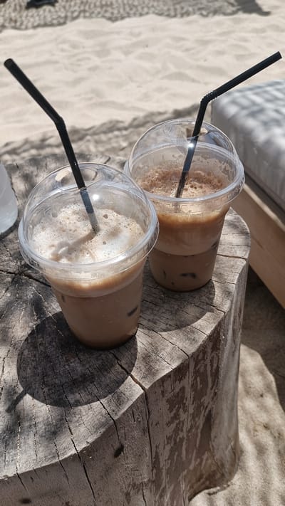 Two iced coffees in clear plastic cups with domed lids and black straws sit on a weathered, circular wooden stump. The coffee in the left cup is lighter in color and topped with foam, while the coffee in the right cup is darker and layered with ice at the bottom. The stump is positioned on a sandy surface, with the texture of the sand visible in the background. In the upper right corner, a portion of a cushioned lounge chair is visible, also casting a shadow. The overall scene suggests an outdoor setting, possibly a beach cafe in Nea Peramos, Greece.