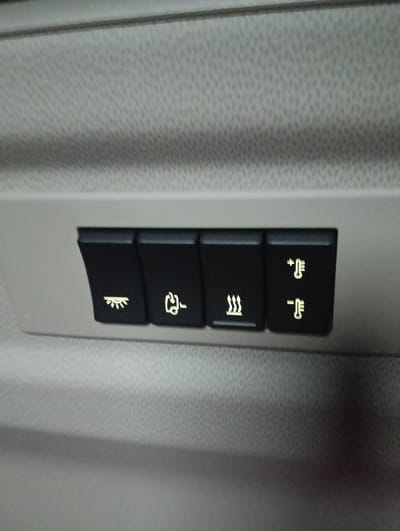 This is a close-up, slightly angled, medium shot of a panel with four illuminated buttons, set within a textured, light-colored surface that appears to be the interior of a vehicle. The buttons are black and rectangular, with the illuminated symbols set within them. From left to right, the symbols depict: a stylized light bulb with rays emanating from it, a handset with a cord and a horizontal line, three wavy lines suggesting heat or steam, and two thermometers, one with a plus sign above it and one with a minus sign above it. The background is an out-of-focus, textured surface. There are no people or overt actions visible. The image is focused on the buttons. The location in Marck, France is not discernible from the image.