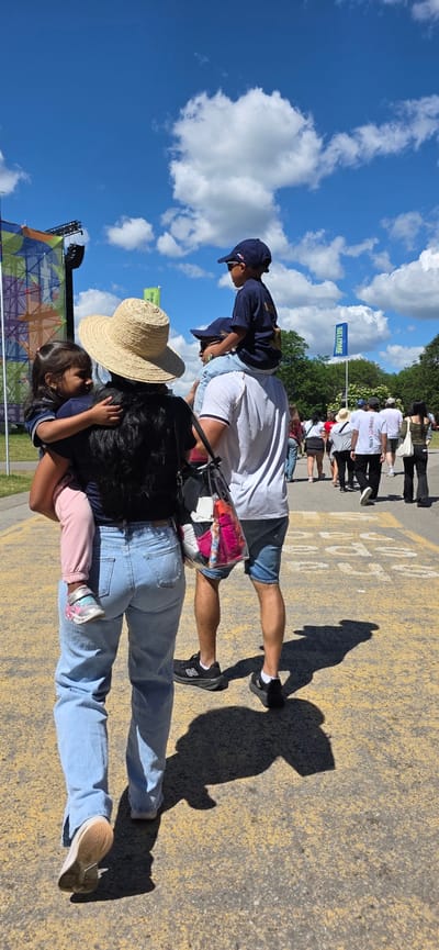 This is a horizontal, eye-level, full-shot photograph taken on a sunny day, likely in the afternoon due to the strong shadows. The scene depicts a group of people walking on a paved surface marked with yellow lines, possibly a road or a large outdoor venue.
In the foreground, a woman wearing a straw hat is carrying a young child on her back. The child is wearing a pink outfit. The woman is dressed in a dark top and blue jeans. Next to her, a man is walking, wearing a white t-shirt, denim shorts, and black sneakers. He is carrying a clear plastic bag filled with clothing. On his shoulders, a young boy wearing a dark blue baseball cap and t-shirt is riding.
In the background, a line of people are walking away from the camera. One person is carrying a white bag. Further in the background, trees line the edge of the paved area. A blue flag with white text that reads "WELCOME" is visible, attached to a pole. To the far left of the image, a large, colorful, geometric structure, possibly a stage or an art installation, is partially visible against a bright blue sky with white clouds.
The overall mood is casual and relaxed, suggesting a family outing or attendance at an outdoor event. The bright sunlight and clear sky indicate pleasant weather. The presence of a "WELCOME" flag and the large structure suggests the location might be a festival, concert, or a public gathering. No other text is clearly legible in the image.
