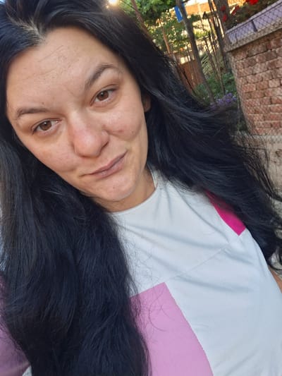 The primary subject is an adult female positioned slightly off-center, gazing directly into the camera. She has long, dark, likely black hair, appearing straight to wavy, which frames her face and extends over her shoulders and chest. Her skin tone is light to medium olive, with visible pores, minor blemishes on her cheeks, and horizontal creases across her forehead. Her eyes are dark brown, and her eyebrows are naturally shaped and dark. Her nose exhibits slight redness at the tip, and her lips are naturally pigmented, forming a subtle, closed-mouth expression. She wears a short-sleeved garment primarily white, featuring a prominent fuchsia or bright pink color-block panel on the right side of her torso (viewer's left). The fabric appears to be cotton or a similar textile, showing some minor textural irregularities.
The scene is set outdoors under direct sunlight, indicated by bright highlights on her hair and the background, suggesting either early morning or late afternoon. The background is partially blurred. To the subject's right (viewer's left), lush green foliage and bright, indistinct light sources are visible. To her left (viewer's right), a weathered red brick wall with visible mortar joints runs horizontally, partially topped by a chain-link fence. Sparse green vegetation, including tree trunks and small clusters of purple flowers, is present behind the fence. Distant, out-of-focus residential structures with reddish roofs and a faint blue sign are discernible in the upper background. The overall environment suggests a residential area or garden in Gotse Delchev, Bulgaria.