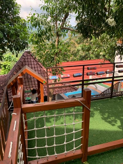 The image shows a wooden playground structure with a rope net. The structure is located on a rooftop with a view of the surrounding forest. There is a small child standing near the edge of the platform wearing a white shirt and blue shorts. The child is looking towards the left of the image. In the background, there is a red roofed building with a metal railing that is covered with various colorful packages. Behind the building, there is a swimming pool that is mostly out of view. There is a green artificial grass surface on the rooftop. The image was taken in Kecamatan Borobudur, Indonesia (latitude: -7.606, longitude: 110.18).