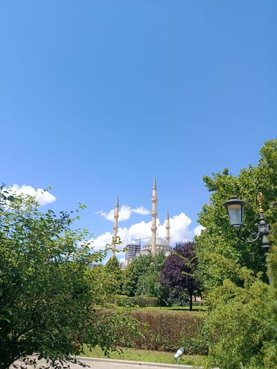 The image shows a bright, sunny day with a clear blue sky and scattered white clouds. In the background, a large, light-colored mosque with multiple minarets dominates the skyline. Construction cranes are visible behind the mosque, suggesting ongoing development. The mosque is surrounded by lush green trees of varying sizes and shades, with some exhibiting a reddish-purple hue. In the foreground, a well-maintained park area is visible, featuring manicured hedges, grassy lawns, and a paved pathway. A vintage-style lamppost with an ornate base stands to the right, partially obscured by foliage. The scene appears to be of the Akhmad Kadyrov Mosque in Grozny, Russia, a prominent landmark in the city. No people are visible in the image.