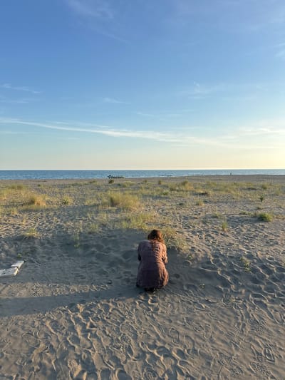 A vertical photograph shows a person on a beach. The sky is blue with sparse clouds. The horizon line divides the image roughly in half, with the sky on the left and the beach on the right. The beach is sandy and littered with footprints and sparse vegetation. The person is wearing a long brown dress and is kneeling on the sand. The image was taken at Donji Štoj, Montenegro.