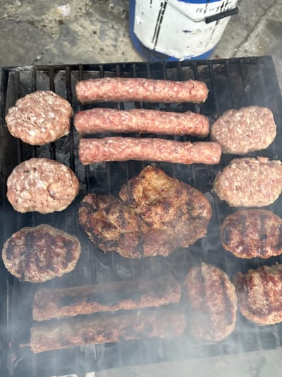 In Kableshkovo, Bulgaria, a close-up, overhead shot captures a barbecue grill laden with various meats. The grill features an assortment of raw and partially cooked items. Three rows of elongated, raw sausages are positioned in the upper section, with raw patties flanking them on either side. Below these, a large, thick cut of meat, showing grill marks and browning, is center-stage. Surrounding this central piece are more oval-shaped patties, some appearing raw and others showing signs of being grilled. The bottom portion of the grill holds two more rows of elongated items, similar to the sausages at the top, but these appear more cooked, with visible browning and charring. Smoke rises from the grill, indicating the active cooking process. The background reveals a concrete or stone surface and the lower portion of a blue and white bucket.