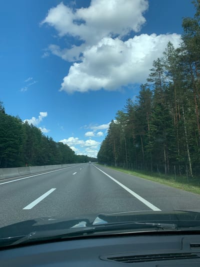 The image shows a view from the driver's seat of a vehicle traveling on a multi-lane highway. The highway is flanked by dense green trees on both sides. The sky is bright blue with scattered white clouds. The road has clear white lane markings. A concrete barrier separates the lanes of traffic. In the distance, a white vehicle is visible on the highway. The foreground shows the dashboard and windshield wipers of the vehicle. The overall scene suggests a sunny day and a journey through a forested area. This location is likely within or near Бакштаўскі сельскі Савет, Belarus, given the visual characteristics of the landscape, which are typical of rural Belarusian highways.