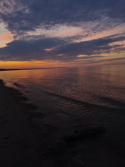 The image depicts a serene sunset over a calm body of water, likely the Baltic Sea, adjacent to a sandy beach. The sky is a canvas of dramatic, dark gray clouds interspersed with soft hues of pink and orange, indicating the last moments of daylight. The water reflects the muted colors of the sky, with gentle ripples disturbing its surface. The foreground features a dark, sandy beach with faint traces of the receding tide. A single, dark-colored piece of driftwood rests partially submerged in the shallow water near the shore. The overall scene is tranquil and slightly melancholic, evoking the quiet beauty of nature at dusk. The location is identified as Apšuciems, Latvia, a coastal village known for its natural beaches and tranquil atmosphere.