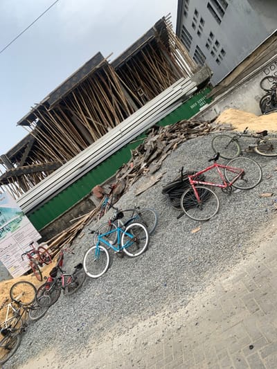 The image depicts an outdoor urban scene in Jakande, Nigeria, under an overcast sky. A multi-story building under construction occupies the upper left and central background, characterized by dark formwork and numerous light-brown wooden scaffolding poles supporting its upper sections. Adjacent to it, on the upper right, stands a multi-story grey building with multiple rectangular window openings.
The foreground is dominated by a large, irregular pile of light grey gravel. Scattered across this gravel pile are approximately ten bicycles in varying states of repair, including a red road bike with black wheels, a light blue bicycle with white rims, and several other dark and mixed-color bikes. A pile of black rubber tires rests near the red bicycle.
Behind the gravel, a green industrial shipping container is visible, marked with the identifier "UACU 57854 45G1" and featuring horizontal metallic reinforcing strips. A substantial heap of dark, broken wooden planks and construction debris is piled on and around the green container. On the far left, a white promotional sign for "THE MERCYPLACE HOUSE" with blue and green graphical elements leans against the gravel pile. The bottom right corner of the foreground displays a section of light brown paved ground, composed of interlocking pavers. No individuals are present in the scene.