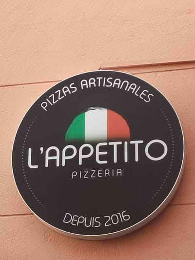 This is a photograph of a circular sign for a pizzeria named L'APPETITO. The sign is mounted on a textured, peach-colored wall. At the top of the sign, curved text reads "PIZZAS ARTISANALES". In the center of the sign, a portion of the Italian flag, featuring green, white, and red vertical stripes, is visible. Below the flag, the name "L'APPETITO" is prominently displayed in large white letters, followed by "PIZZERIA" in smaller white letters. At the bottom of the sign, the text "DEPUIS 2016" indicates the establishment's founding year. The sign has a metallic rim and a dotted border around the inner edge. The lighting suggests it is daytime, and the overall scene is a straightforward depiction of a business sign. The context provided indicates the location is Perpignan, France.