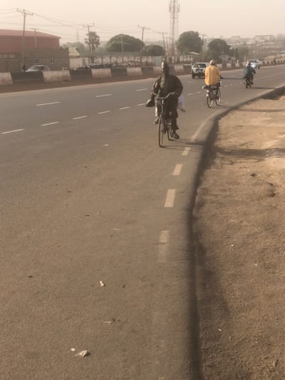 The image depicts a daytime street scene on an asphalt road in Hanwa, Nigeria, under a hazy, light-grey sky. The road surface is light brown-grey with scattered debris. White dashed lines delineate the right edge of the paved road, which transitions to an unpaved dirt shoulder on the right.
In the foreground, a male individual dressed in dark clothing (possibly a dark agbada or similar garment) rides a dark-colored bicycle. He carries a white object, possibly a bag, on the front of his bicycle, and a dark bag or garment over his left shoulder. He faces forward, moving away from the viewer at a slight angle. Long shadows are cast from the right side of the frame onto the road surface.
Further down the road, a second male individual, wearing a bright yellow long-sleeved shirt and dark trousers, rides a bicycle away from the viewer. A white item is secured to the rear rack of his bicycle. Behind him, a third individual rides a motorcycle, also moving away. In the distant background, several light-colored cars are visible on the multi-lane road.
The background features a concrete barrier separating the road from various structures. To the left, a large, reddish-brown building with a flat roof is visible. Beyond the barrier, several smaller buildings with white walls and dark roofs are present, along with green trees and utility poles. A tall, grey metal telecommunications tower stands prominently against the hazy sky in the far background.
