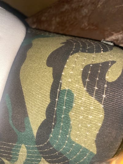 The image shows a close-up, angled view of a camouflage-patterned fabric, likely from a hat or clothing item. The pattern consists of irregular shapes in shades of green, black, and khaki. Visible stitching runs in parallel lines across the fabric. A white fabric is partially visible on the left side, and a blurred, brown, textured background is present in the upper right corner. The scene is indoors and appears to be a still life, with no people or actions depicted. The location in Tavo, Nigeria, is not evident from the visual information in the image.