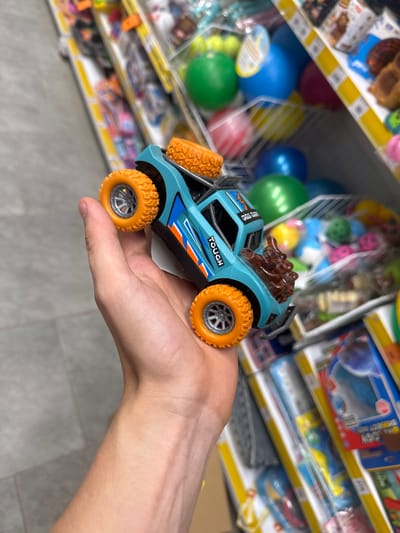 A person is holding a blue and orange toy monster truck in their hand. The toy truck has large orange tires and the word "TOUGH" is visible on its side. In the background, shelves are stocked with various toys, including colorful balls. The scene appears to be in a store. The overall lighting is bright, suggesting an indoor environment. There is no visual information in the image to indicate the location as Ivanivka, Ukraine.