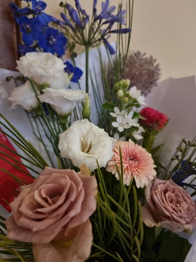 The image shows a close-up of a floral arrangement. The bouquet features a variety of flowers, including muted pink roses, white lisianthus, a pink gerbera daisy, blue delphiniums, and small white flowers. The arrangement is presented against a neutral, light-colored background, likely an indoor setting. There is no indication of a specific location in Givatayim, Israel, or any people or actions. The focus is solely on the detailed textures and colors of the flowers.