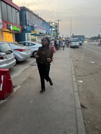 A woman in a floral shirt and black pants walks on a sidewalk, looking at the camera. Parked cars flank her, and a "Pinkberry" restaurant is partially visible. Behind the cars, businesses line the street. In the distance, traffic moves along a road, and a cloudy sky is overhead. The scene has a slightly tilted perspective.