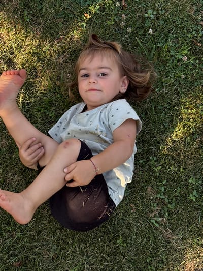 A young girl with light brown hair lies on her back in a grassy area. She is wearing a light blue t-shirt with small white stars and black shorts. Her right arm is bent at the elbow and her hand is resting on her thigh. A thin red bracelet with a four-petal flower charm is visible on her wrist. She has a small abrasion on her right knee and elbow. Her left leg is bent at the knee, and her left foot is visible in the lower left corner of the frame. The girl is looking up and to her left, with a slight smile on her face.
The scene appears to be outdoors in a park or yard, with dappled sunlight suggesting it is daytime, likely late afternoon due to the angle of the light. The grass is green and appears healthy, with a few small white flowers scattered throughout. The setting is likely Oreshak, Bulgaria. There is no visible text in the image. The overall mood is relaxed and carefree.