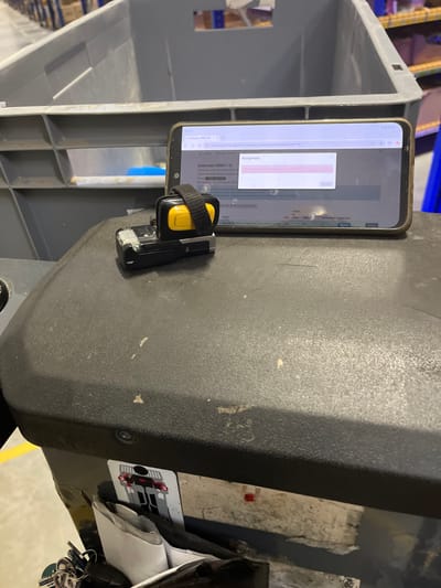 The image depicts a close-up view of a black textured surface, likely the dashboard of a vehicle or a piece of industrial equipment, in Tarnowo Podgórne, Poland. A smartphone is propped up, displaying a webpage with Polish text. A small device with a yellow button and a black strap is attached to the smartphone. The background shows large grey plastic bins, commonly used in warehouses or logistics facilities. There are also some keys and what appears to be a white paper object visible in the lower part of the image. The scene suggests a work environment, possibly related to inventory management or logistics.