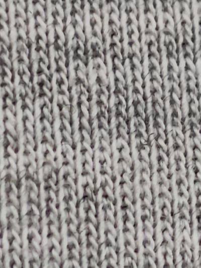 The image is a close-up, macro photograph of a grey, knitted fabric. The knit pattern features vertical ribs formed by interlocking loops of yarn. The yarn appears to be a blend of white and grey fibers, creating a marled or heathered effect. The texture is slightly fuzzy, indicative of wool or a wool blend. There are no people or discernible objects other than the fabric itself. The lighting is even, highlighting the texture and details of the knit. The background is entirely the knitted material. There is no information about the location in Svietlahorsk, Belarus, or any context beyond the visual content of the fabric.