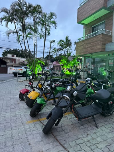 The image shows a row of parked electric scooters on a paved sidewalk in Florianópolis, Brazil. The scooters are various colors, including red, yellow, and green. A chain secures the scooters together. In the background, palm trees line the street, and buildings are visible, one with green tinted lighting. The sky is overcast. A white car is parked behind the scooters. The address "5" is visible on one of the buildings.