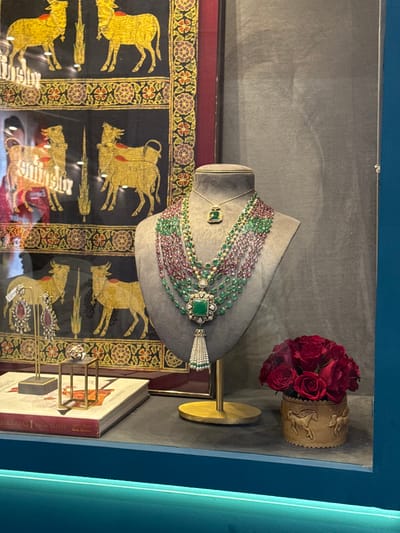 This image depicts a high-end jewelry display within a retail environment located in the city of Jaipur Municipal Corporation, India. The central subject is a gray mannequin bust showcasing an elaborate multi-strand necklace composed of numerous strands of polished green emerald beads and reddish-pink faceted beads. This necklace features a large, square-cut emerald pendant surrounded by smaller white stones, from which a white pearl tassel descends. A shorter golden necklace with a smaller, rectangular emerald pendant hangs above the main piece.
To the left of the mannequin, a pair of ornate dangle earrings, set with large red oval gemstones encircled by smaller white stones, rests on a slender golden stand. Next to the earrings, a ring with red and white stones is displayed on a small, geometric gold-toned stand. Beneath these items, a stack of books is visible, with the top volume titled "MAHARAJAS AMIN JAFFER MARK BORTHWICK."
To the right of the mannequin, a small, cylindrical light-brown container embossed with horse figures holds a compact bouquet of deep red roses. The entire display is situated on a dark, flat shelf, with a horizontal strip of teal-colored illumination along its front lower edge.
In the background, behind the jewelry, a large framed textile artwork or painting features four stylized golden cattle, likely cows or bulls, against a black background, framed by an intricate decorative border. Reflections, including partial human forms, are visible on the clear glass surface enclosing the display, indicating a shop window or display case. The background wall behind the framed art is a uniform dark gray.