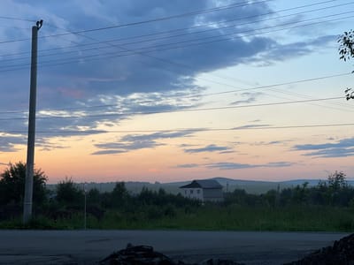 The image depicts a serene rural landscape at dusk, located in the city of Druzhinino, Russia. The sky transitions from a deep blue with scattered clouds at the top to hues of orange and pink near the horizon, indicating the setting sun. A prominent utility pole with multiple overhead wires stands on the left side of the frame, positioned near a paved road. A speed limit sign, displaying "40" within a red circle, is visible on a post to the right of the utility pole. In the middle ground, a two-story house with a dark roof is nestled amongst dense green foliage and trees. The background features rolling hills and fields, with scattered lights suggesting a distant settlement. The foreground consists of a dark, rough surface, possibly a road under construction or debris. No people are present in the image.