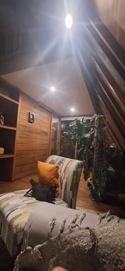 The image captures an indoor scene in what appears to be a loft or attic space with a sloped wooden ceiling. A bright overhead light fixture casts strong rays. The room is furnished with a striped chaise lounge and a patterned armchair, both with cushions. A black bag rests on the chaise lounge, next to an orange cushion. A grey throw blanket with fringe is draped over the foreground of the chaise. To the left, a wooden shelf unit is visible, holding a decorative item. In the background, a glass door leads to an area with large green plants. The walls are paneled with wood. The location is identified as Banjar, Indonesia.