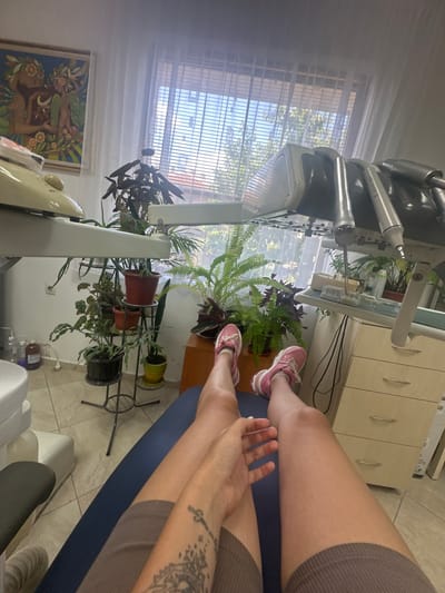 A person's legs are visible, extending into the frame from the bottom left, as they lie on a blue dental chair. The person is wearing brown shorts and pink sneakers. To the left of the dental chair, a white dental unit is partially visible. In the background, there are several potted plants, a window with blinds, and a framed artwork on the wall. A dental cabinet with drawers is situated to the right. The scene appears to be inside a dental clinic.