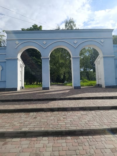 The image shows a light blue archway with three openings, set against a background of trees and a cloudy sky. The archway is constructed of concrete and painted white and blue, with decorative diamond shapes above each arch. In front of the archway is a paved walkway made of interlocking bricks, with three steps leading up to the structure. Beyond the archway, a path leads into a park-like area with lush green grass and trees. The overall impression is one of an entrance to a park or public space. This archway is located in the city of Khorol, Ukraine.