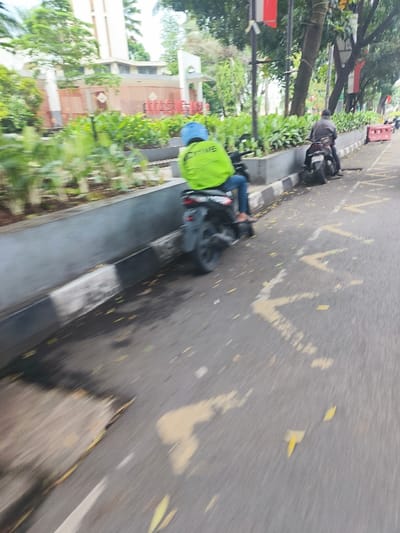 The image shows two motorbikes on the side of a road in Cimahi, Indonesia, at the coordinates -6.873 latitude and 107.541 longitude. The first motorbike is stopped on the right side of the road, and the rider is wearing a black helmet and a dark jacket. The second motorbike is in motion, and the rider is wearing a blue helmet and a green jacket with the word "Drive" written on it. The rider is moving forward, with the back of the motorbike and the rider's legs visible. Both motorbikes are facing the same direction, and the road is relatively empty with a few leaves scattered on the asphalt. The sidewalk is lined with a curb and a row of green bushes, and there are tall trees in the background.  There is a gray concrete wall on the left side of the road, partially obscured by the bushes. There are several red flags on the poles in the background.  The image seems to be taken from the perspective of someone following behind the second motorbike.