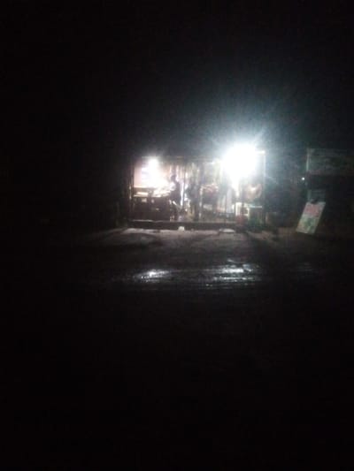 This image depicts a nighttime scene in front of a brightly lit shop, possibly in Television, Nigeria. The shop appears to be a small, open-air establishment with goods displayed on shelves and tables. The light source is very intense, creating a glare that obscures some details. A person, whose features are indistinct due to the lighting, is visible inside the shop. The ground in front of the shop is wet, reflecting the light and suggesting recent rain. The surrounding area is dark, indicating it is nighttime and possibly a less developed part of the city. A sign is visible on the right side of the image, partially illuminated. The overall impression is one of a humble, active commercial space operating after dark.