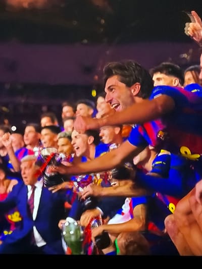 A crowd of people wearing blue and red clothing is gathered, appearing to celebrate something. They are holding a bottle of alcohol and some of them have confetti. They look happy and excited. The photo is taken in Sant Martí Sarroca, Spain.