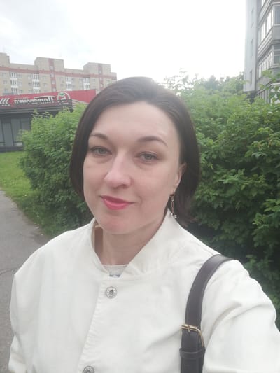 A medium shot captures a woman with dark brown hair, fair skin, and blue eyes, wearing a white jacket and a dark strap over her shoulder, in an outdoor setting. She has makeup on, including pink lipstick and eye shadow. A dangling gold earring with silver accents is visible on her right ear. Behind her, a dense green hedge forms a backdrop. To the left of the hedge, a paved walkway is visible, leading towards a building complex with multiple stories. The building on the left is light brown with red accents and windows, and a red sign with white lettering that reads "Пятерочка" (Pyaterochka), a Russian supermarket chain. To the right of the hedge is a tall, grey building with multiple windows. The sky is overcast. The scene appears to be in the city of Tosno, Russia.