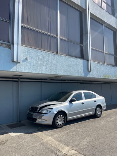 A silver sedan with a damaged front end is parked in front of a blue and gray building. The car's hood is crumpled and the front bumper appears to be damaged. The building has large windows with vertical blinds and a textured blue facade on the lower level. The parking area is paved with asphalt and concrete. The scene appears to be in an urban environment, possibly an industrial or commercial area. There is no graffiti visible on the building in the provided crops. The image is captured from a slightly low angle, looking up towards the building and the car. Sunlight is casting shadows, indicating a clear day. The overall impression is one of neglect or disuse, suggested by the damaged vehicle and the somewhat weathered appearance of the building.