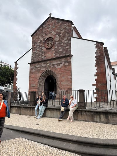 The image depicts a view of the Igreja do Colégio (Church of the College) in Funchal, Portugal. The church's facade is characterized by alternating panels of white stucco and reddish-brown brickwork. A large arched entrance is visible, as is a circular window above the door. The building is situated on a paved street composed of small, patterned tiles. Several tourists are present in front of the church, including people standing near a black metal railing and others walking along the sidewalk. The weather appears to be overcast.