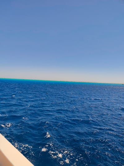 The image depicts a vast expanse of the Red Sea under a clear, bright blue sky. The water is a deep, rich blue, with small waves creating white foam and ripples on the surface. In the distance, a narrow strip of turquoise water signifies a coral reef, with a lighter sandy coastline visible beyond it. The horizon line is sharply defined, separating the sea from the sky. In the foreground, the edge of a light-colored boat railing cuts across the bottom left corner, indicating the perspective is from a vessel on the water. The scene is devoid of any human figures or specific man-made structures, except for the implied presence of the boat. This view is consistent with the coastal scenery of Al Hadaba, Egypt, a popular tourist area known for its clear waters and coral reefs.