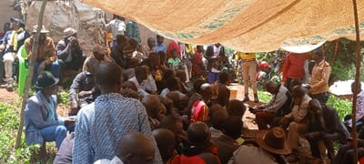 The image depicts a crowded outdoor gathering in Kundu 1, Democratic Republic of the Congo. Approximately 50 to 70 individuals, primarily adult males, are assembled under a large, light-colored tarp or canopy, possibly made from woven material with green markings, supported by rough wooden poles. Most attendees are seated directly on the reddish-brown earth ground, with some occupying a green plastic chair on the far left or resting on low, unseen structures.

Key figures include a man in the left foreground wearing a light blue suit jacket, blue jeans, and a black bowler hat, holding a small brown bottle. Another man in the immediate foreground, with his back to the viewer, wears a black and white patterned shirt. Various men throughout the scene wear casual clothing, including t-shirts, button-down shirts, and trousers; several wear hats such as baseball caps and fedoras. A few women and children are also visible among the seated crowd.

In the background, more individuals are standing, including one person in a yellow top and light-colored pants on the right, and another in a red shirt further left. The crowd appears attentive, with many individuals facing generally towards the right, suggesting a common point of focus or speaker. The environment features sparse green vegetation and dense foliage in the background, with the canopy providing shade from direct sunlight. No specific aggressive actions or overt interactions are discernible beyond general congregating.