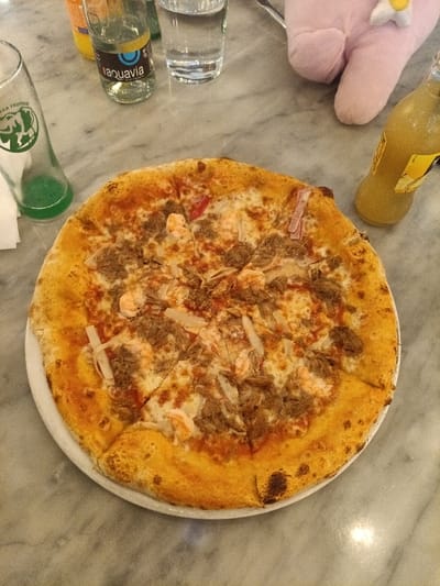 A pizza pie sits on a white plate, cut into uneven slices. It has a golden-brown crust with toppings including what appears to be meat, cheese, and shrimp. A glass of green liquid, a bottle of sparkling water, a glass of water, and a yellow bottle are nearby. A pink plush toy is present in the upper right corner of the image. The surface is a marble or stone countertop.