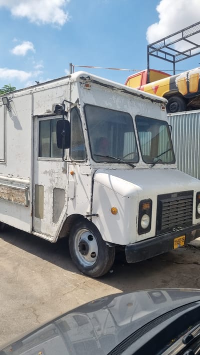 A white, older model delivery truck is parked on a concrete surface in what appears to be an outdoor industrial or storage area. The truck is white with peeling paint, revealing rust and primer underneath. It has a large, flat windshield, a visible driver's side mirror, and a large grille on the front. A license plate with yellow characters on a black background is visible on the front bumper. To the right of the truck, a corrugated metal fence runs along the background. Behind the truck, another vehicle, possibly a truck or bus, with a yellow and red paint scheme and a graphic of beer mugs, is partially visible, resting on a metal structure. The sky above is blue with scattered white clouds. The location is identified as Kfar Saba, Israel.