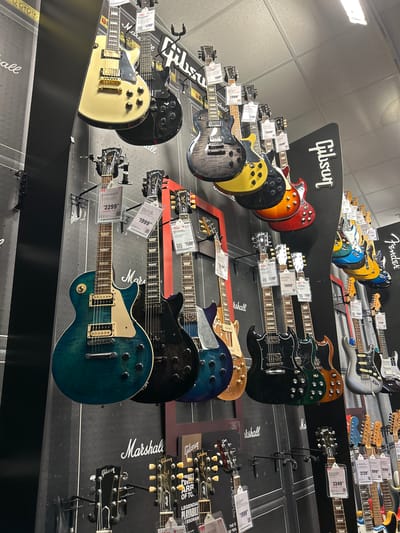 The photograph appears to be taken inside a music store. The main subjects are numerous electric guitars displayed on a wall. The guitars are various models and colors, including brands like Gibson and Fender, with price tags attached to some. The display wall is branded with the logos of Gibson and Marshall. The guitars are hanging on a black wall display. The prices of some of the guitars visible range from $1599 to $2999. Based on the presence of the "Fort Myers" tag and Gibson and Fender Guitars, this is likely a retail music store located in Fort Myers, Florida, USA.